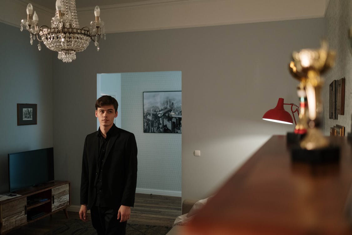 A man in a black suit standing in a dimly lit room with a chandelier and desk lamp