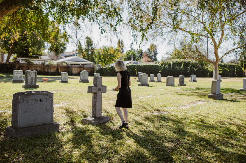 woman in black dress in a cemetery approaching a graves headstone with flowers