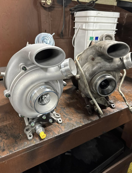 Two turbochargers sit side-by-side; one new and silver, the other older and dirtier, on a workbench.