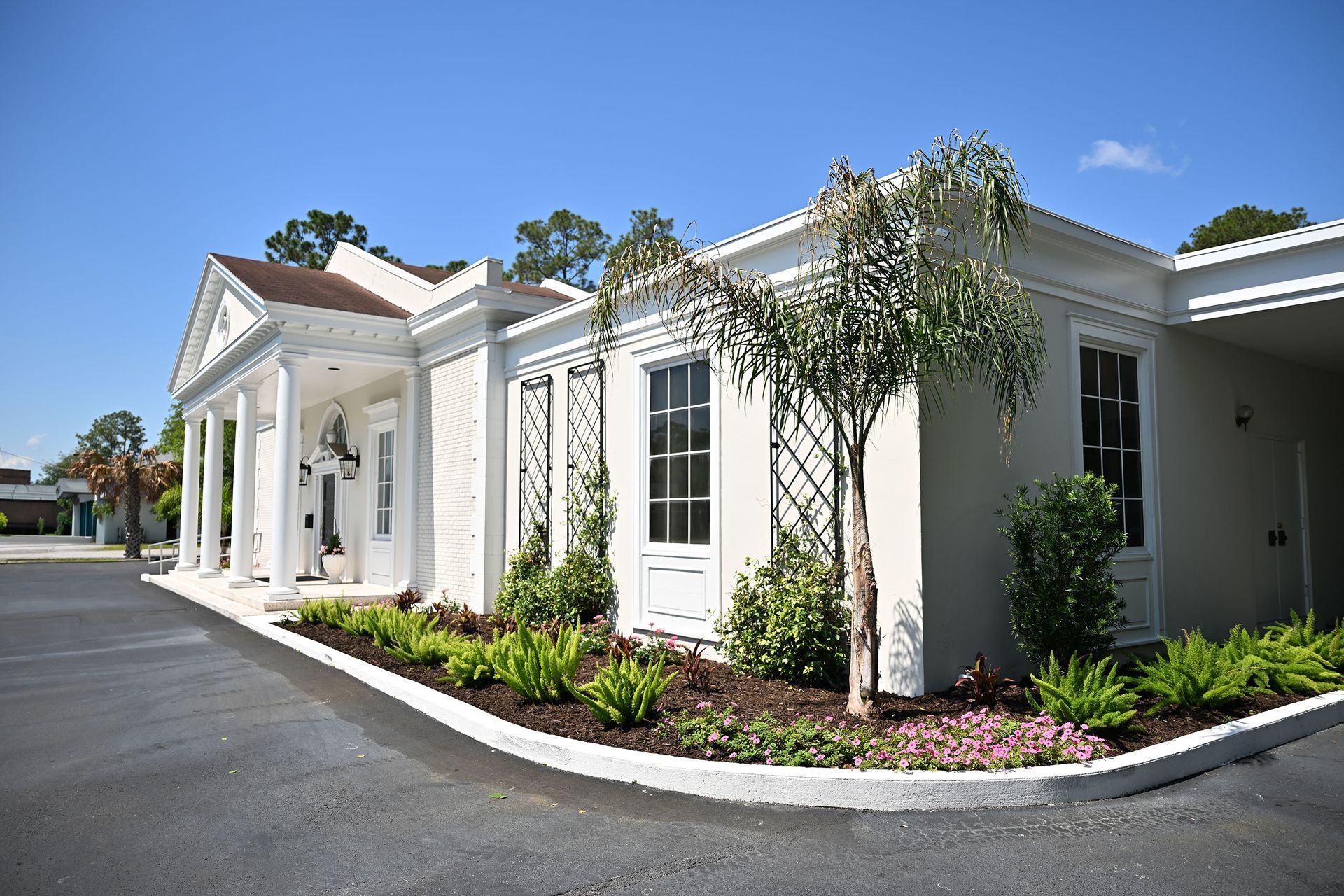 White single-story building with columns, windows, and landscaped shrubs beside a paved driveway