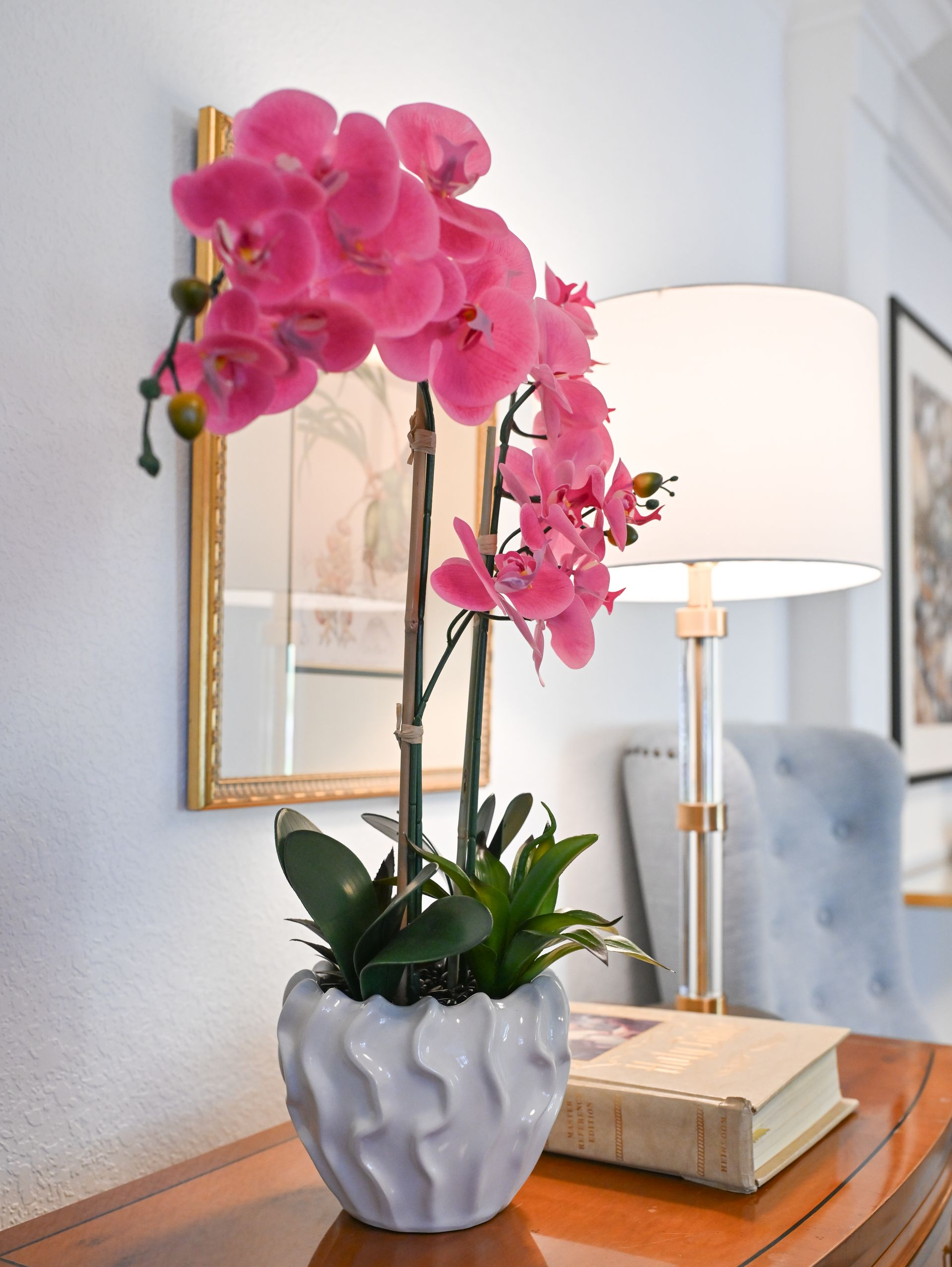 Pink orchid in a white pot on a table beside a book, with a lamp and mirror in the background.
