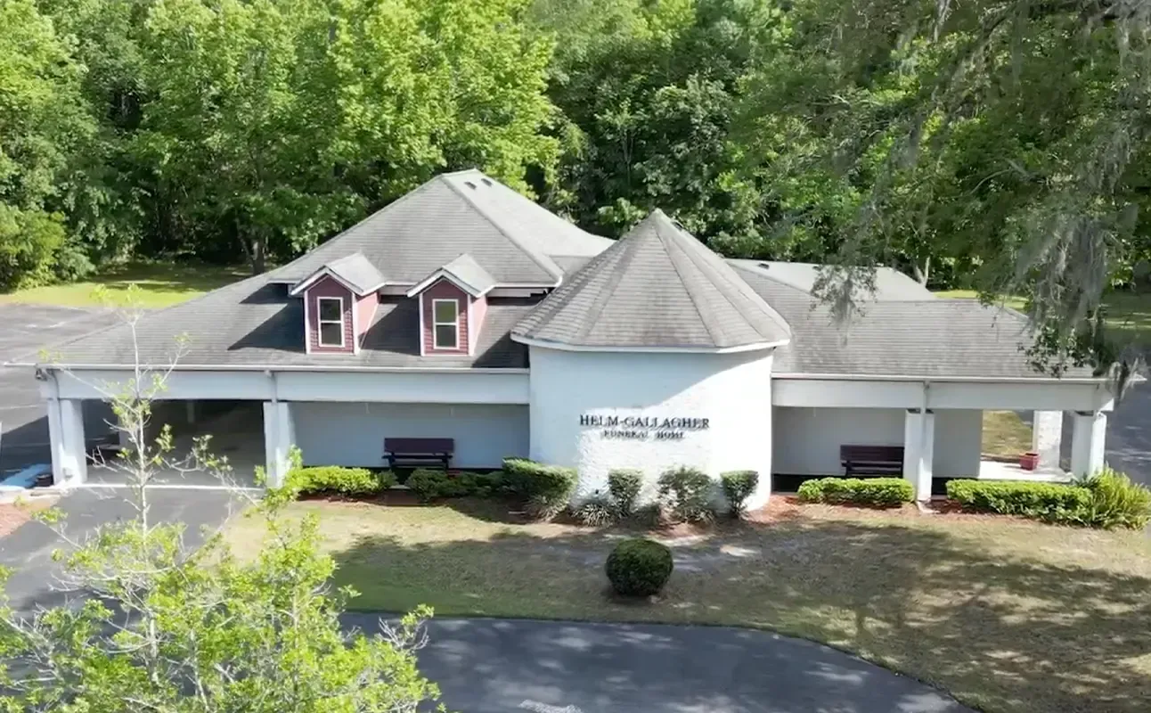 Front view of a white building with a gray roof and trees behind it, with a circular driveway in front