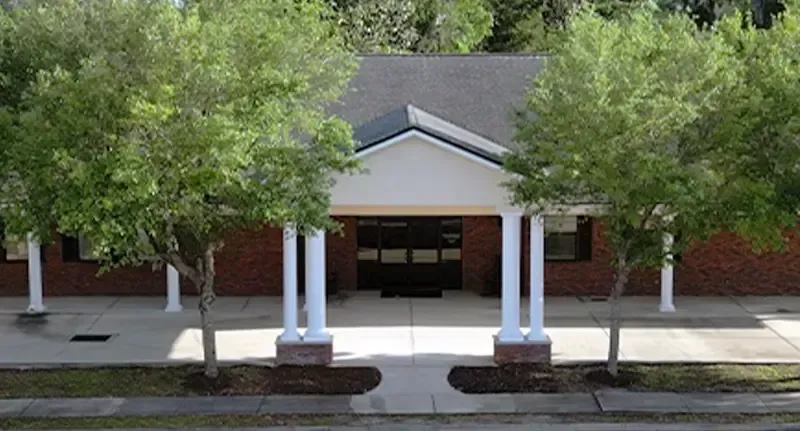 Front entrance of a brick building with white columns, centered walkway, and trees on both sides