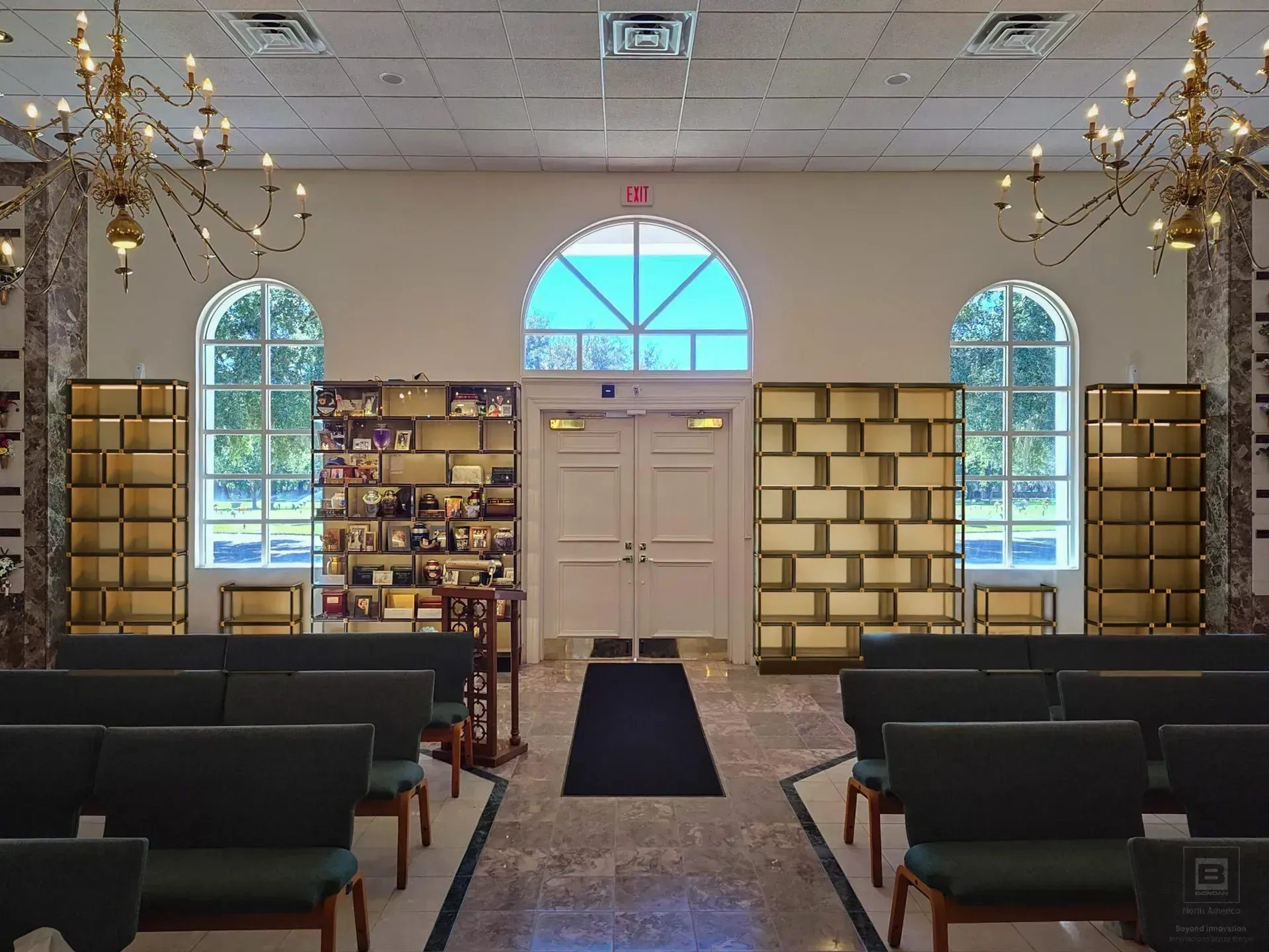 Interior of a small chapel or prayer room with pews, windows, and a central door under a blue arch.