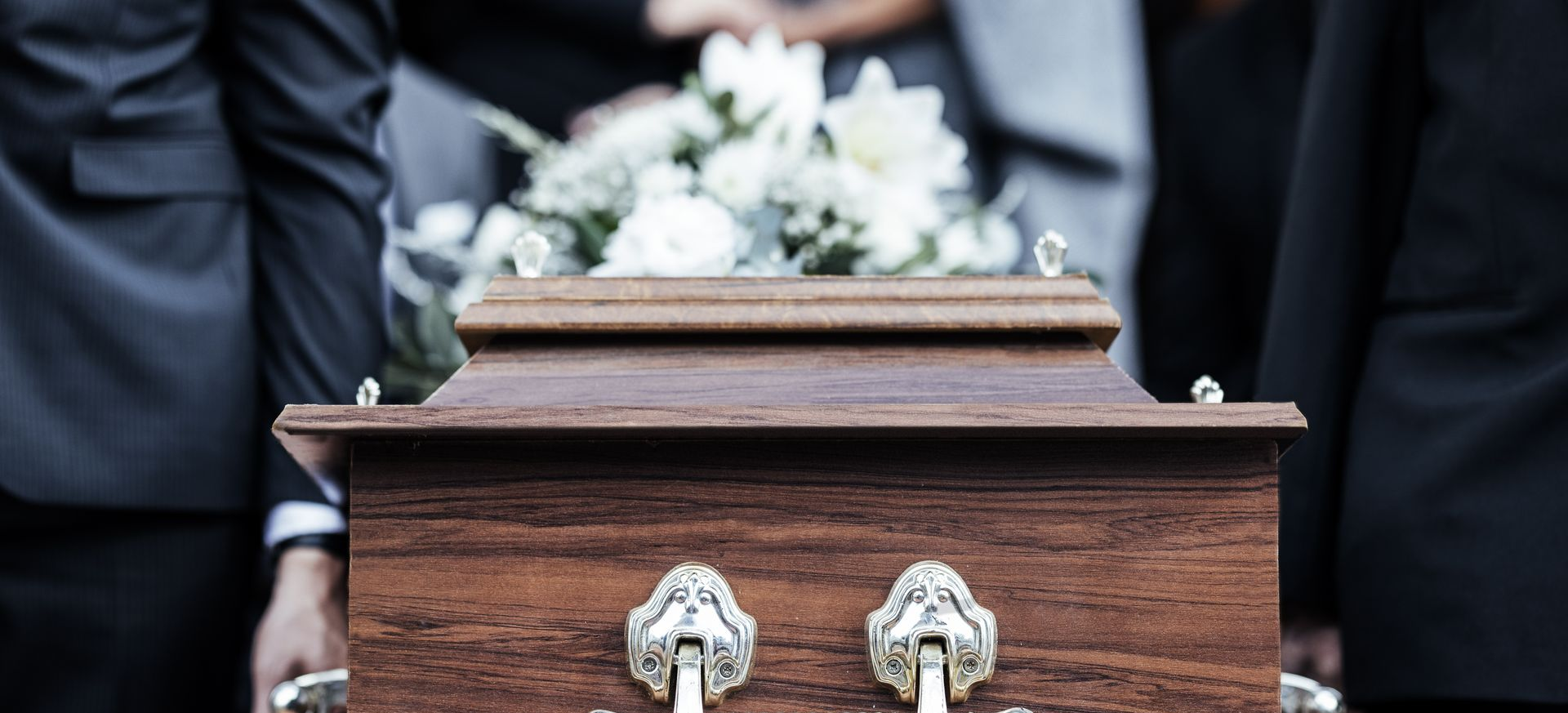 Wooden casket at a funeral, with blurred mourners in dark suits behind it