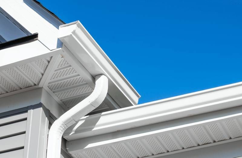 White rain gutters and downspout on a house with gray siding against a bright blue sky.