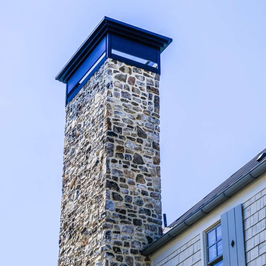 Stone chimney on a building with a dark, square top against a clear sky.