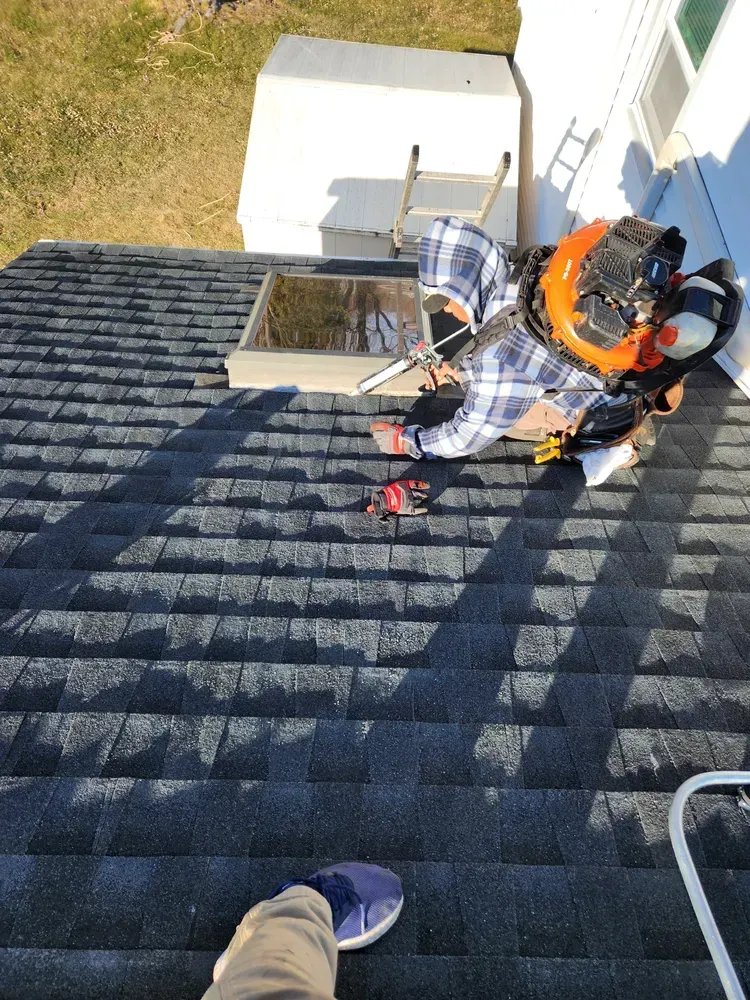 Person on a roof working near a skylight, ladder nearby. Sunny day, grey shingles.