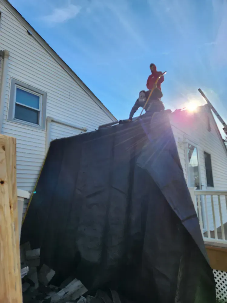 Two people on a roof covered in black tarp, working on a house with white siding under a sunny sky.