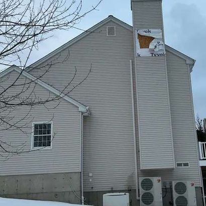 Side of a light-colored house with exposed Tyvek wrap on the chimney and two HVAC units.