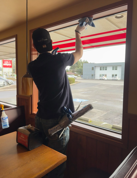 Person cleaning a large window in a restaurant, using a squeegee and cloth, with a view of a parking lot.