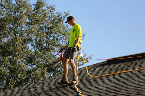 Roofer wearing safety harness stands on a roof, holding a tool. Bright shirt, blue sky.