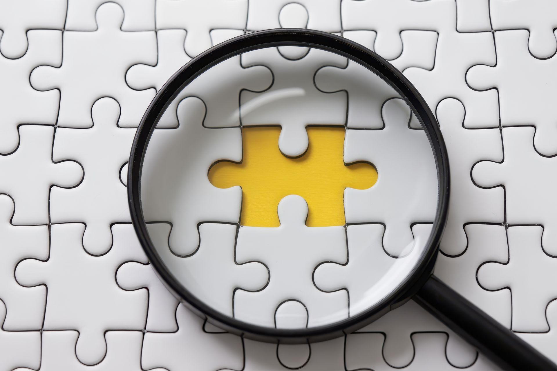 Magnifying glass over missing yellow puzzle piece in a white puzzle.