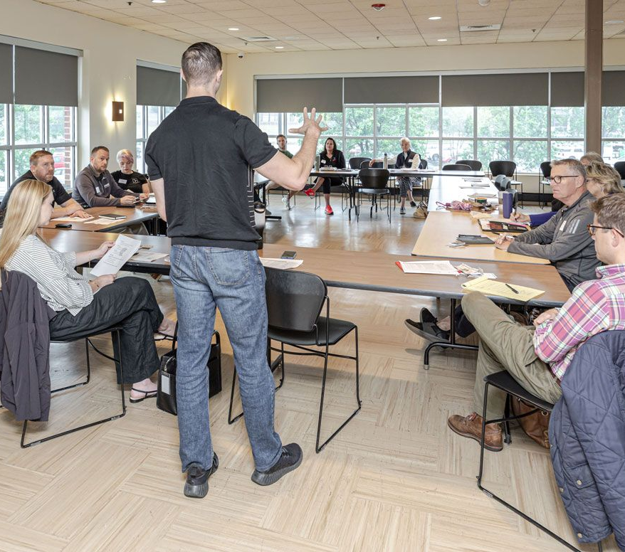 Teacher leading a classroom discussion with students seated around tables in a bright room
