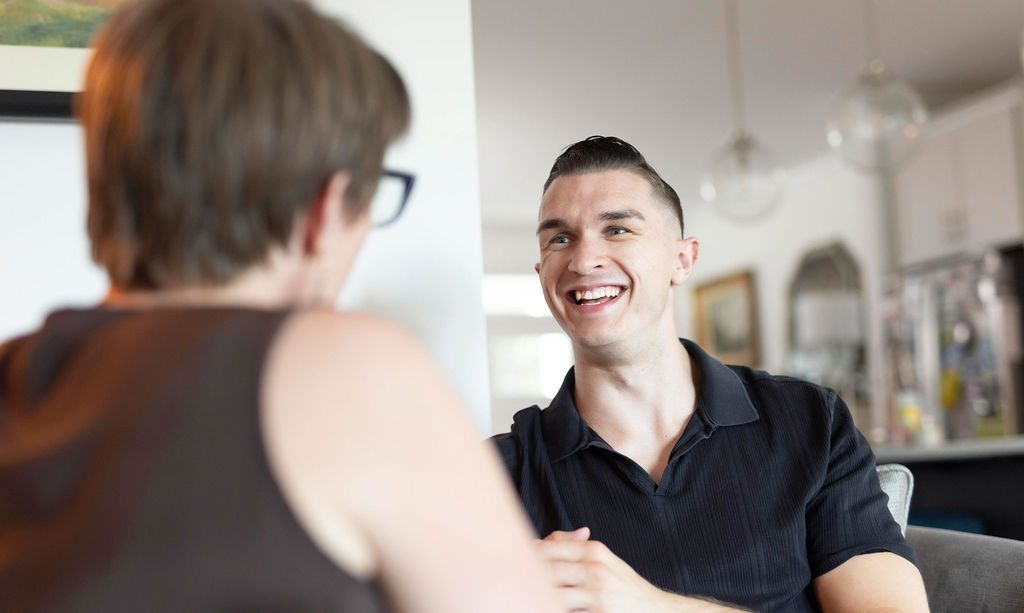 Man in black shirt smiles at person seated opposite; indoor setting, bright lighting.