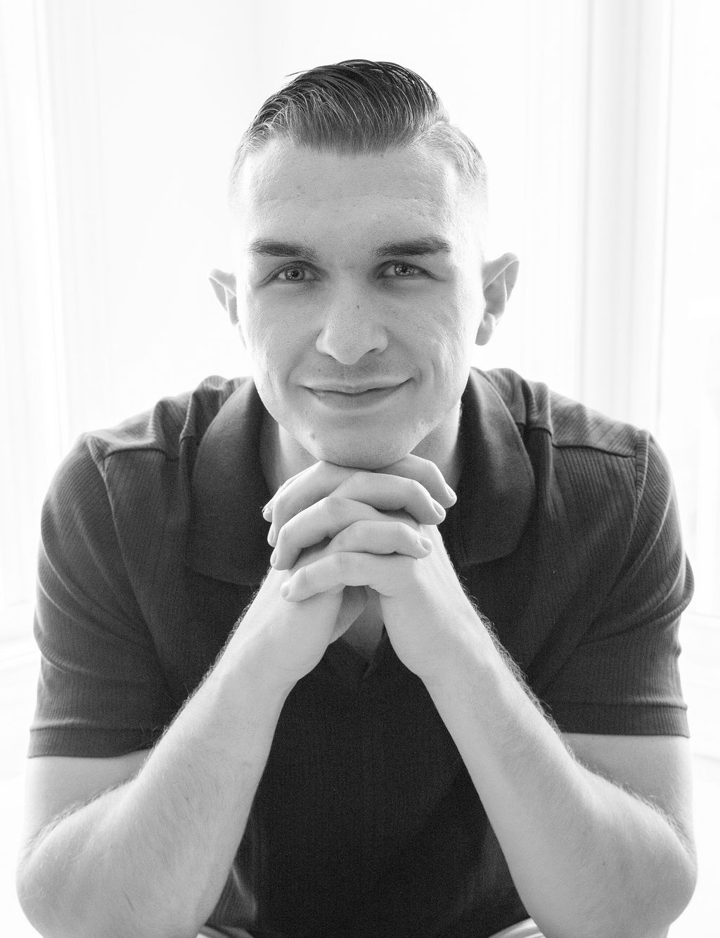 Man in dark shirt, clasped hands under chin, smiles at camera. Soft, natural light.