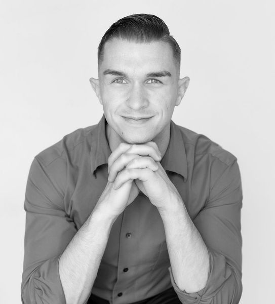 Man in red shirt, hands clasped under chin, smiling against a white backdrop.