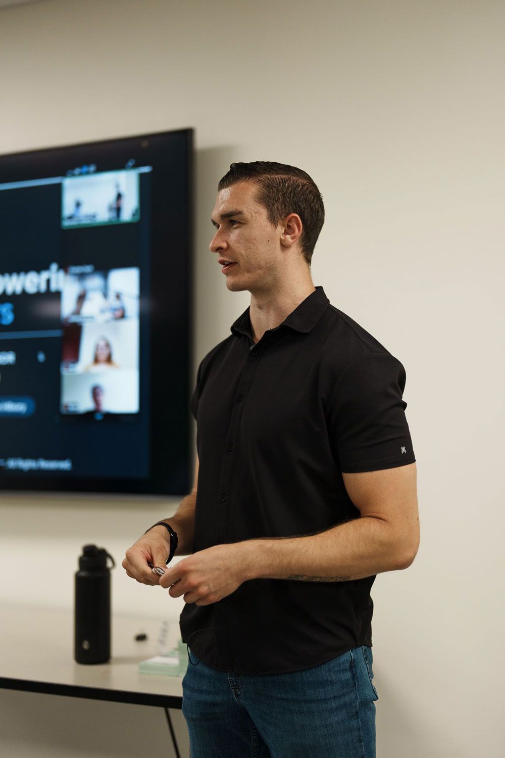 Man speaking beside a wall-mounted screen in a meeting room, wearing a black polo and jeans.