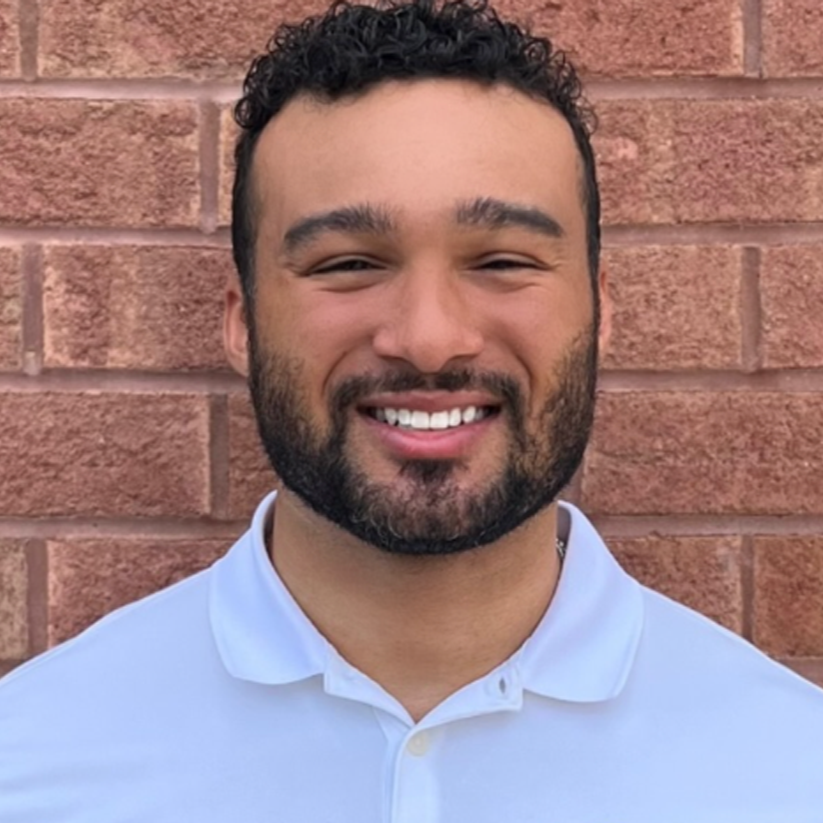 Man with a beard smiling in front of a brick wall, wearing a white polo shirt.