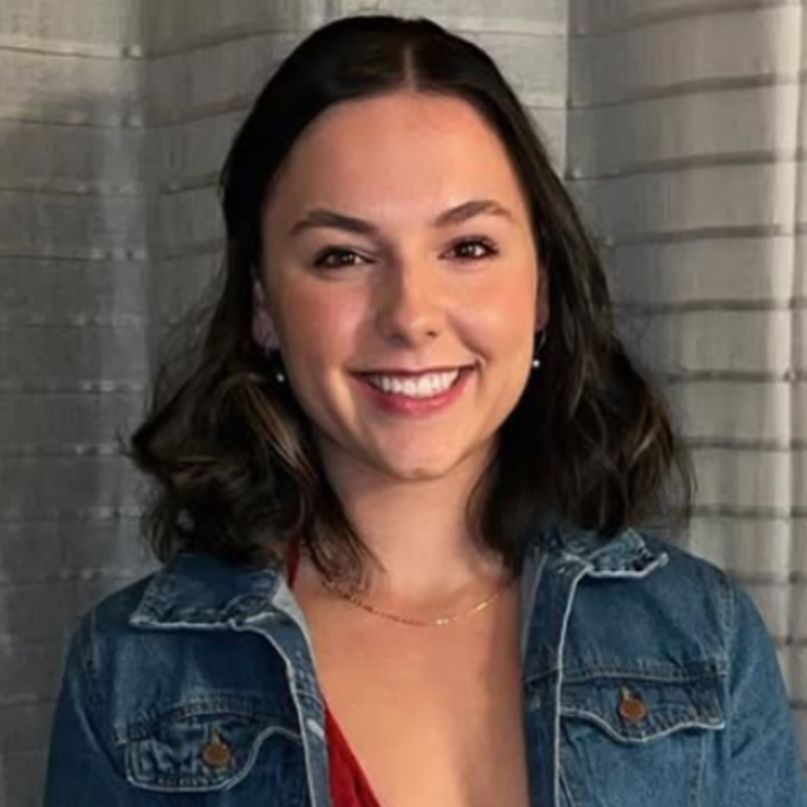 Woman smiling, wearing a denim jacket and a red top, in front of a window with blinds.