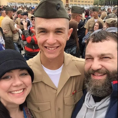 A young man in a Marine uniform smiles with two people. Outdoor setting, other people in background.