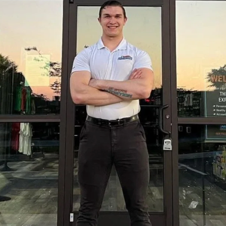 Man in white shirt and dark pants, arms crossed, standing in front of glass doors.