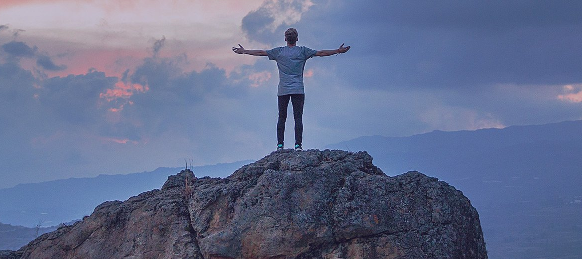 Person standing on a rocky peak with arms outstretched at sunrise over mountains