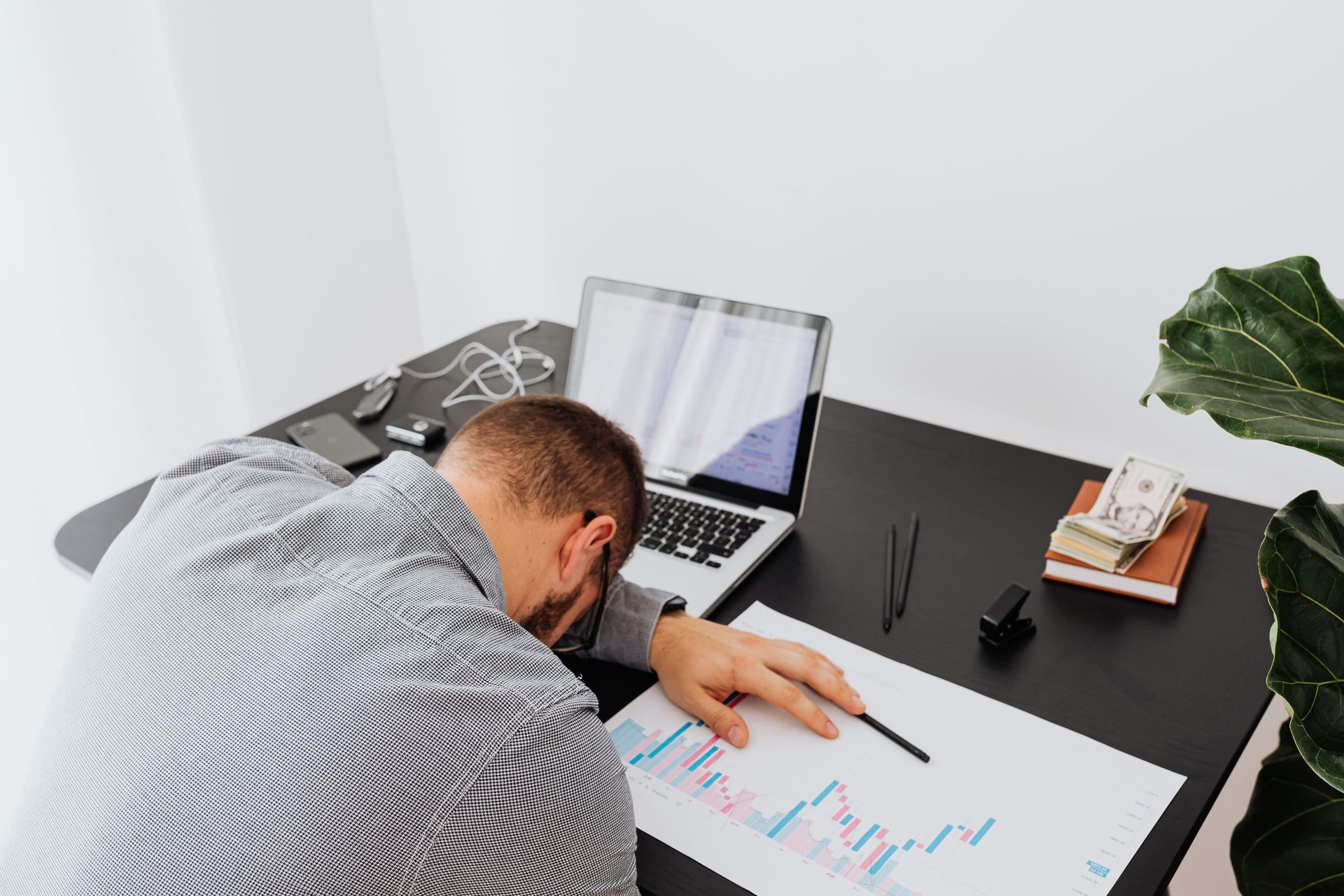Person leaning over a desk, reviewing charts beside a laptop and plant.