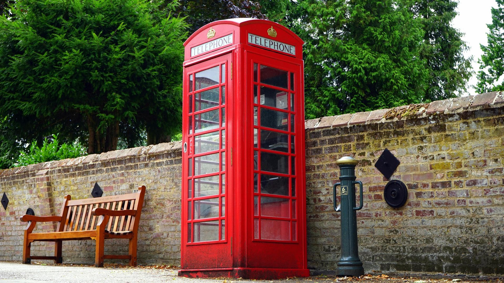 Red British phone booth beside a wooden bench and stone wall, with green trees in the background.