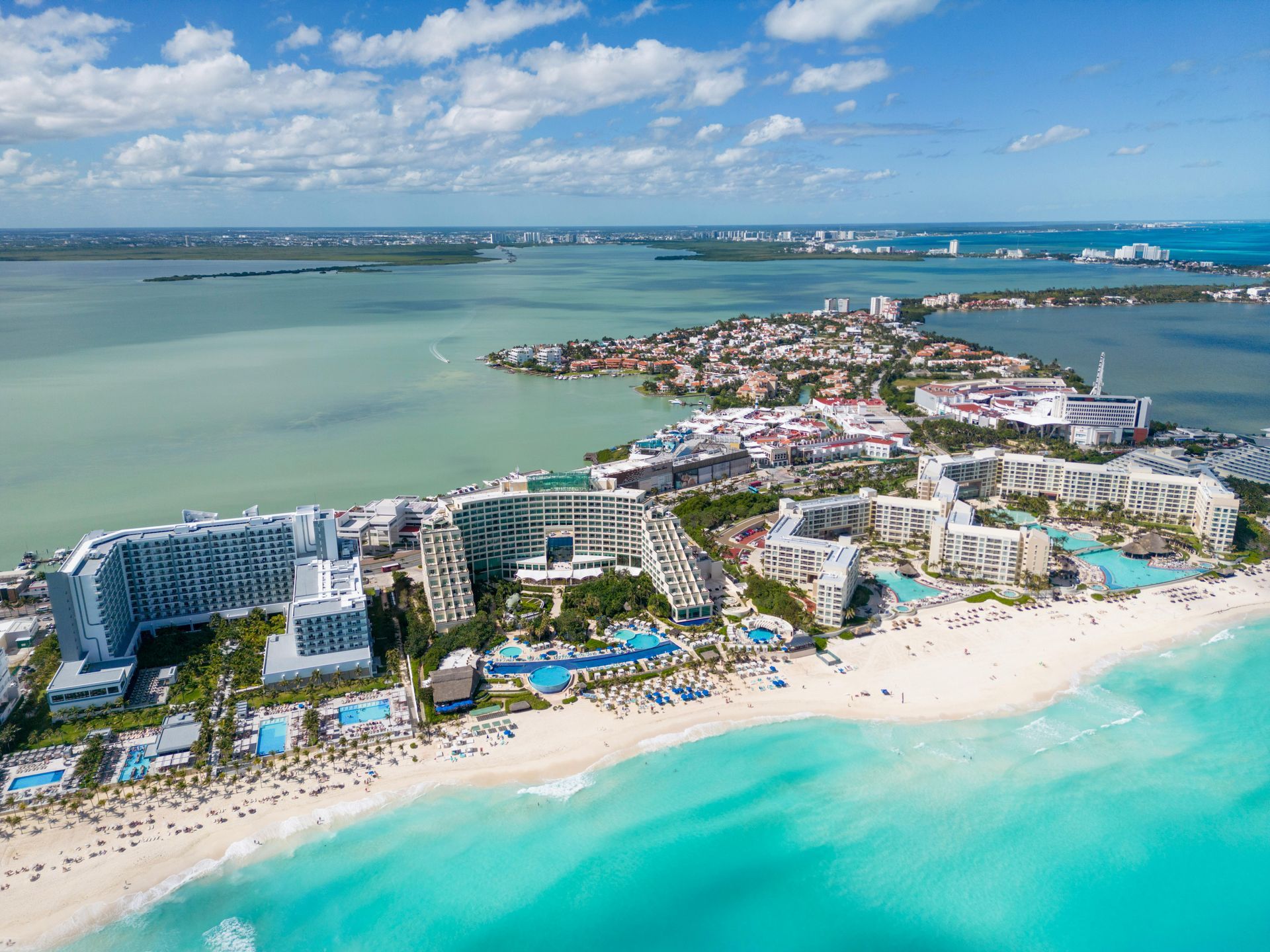 Aerial view of a turquoise beach resort with white buildings, pools, and a sandy shore near a small island