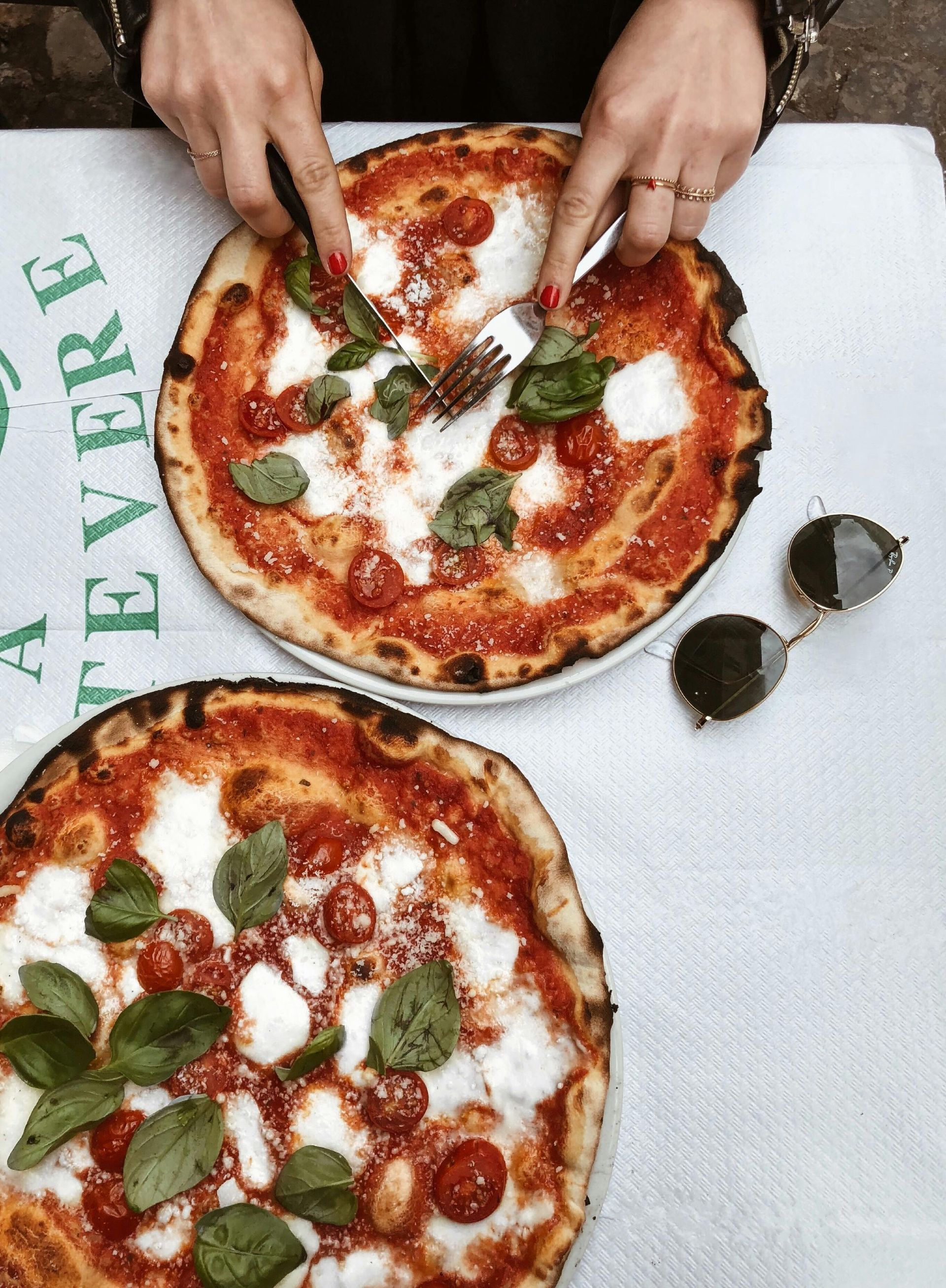 Hands placing basil on two cheese pizzas with pepperoni on a white table beside sunglasses