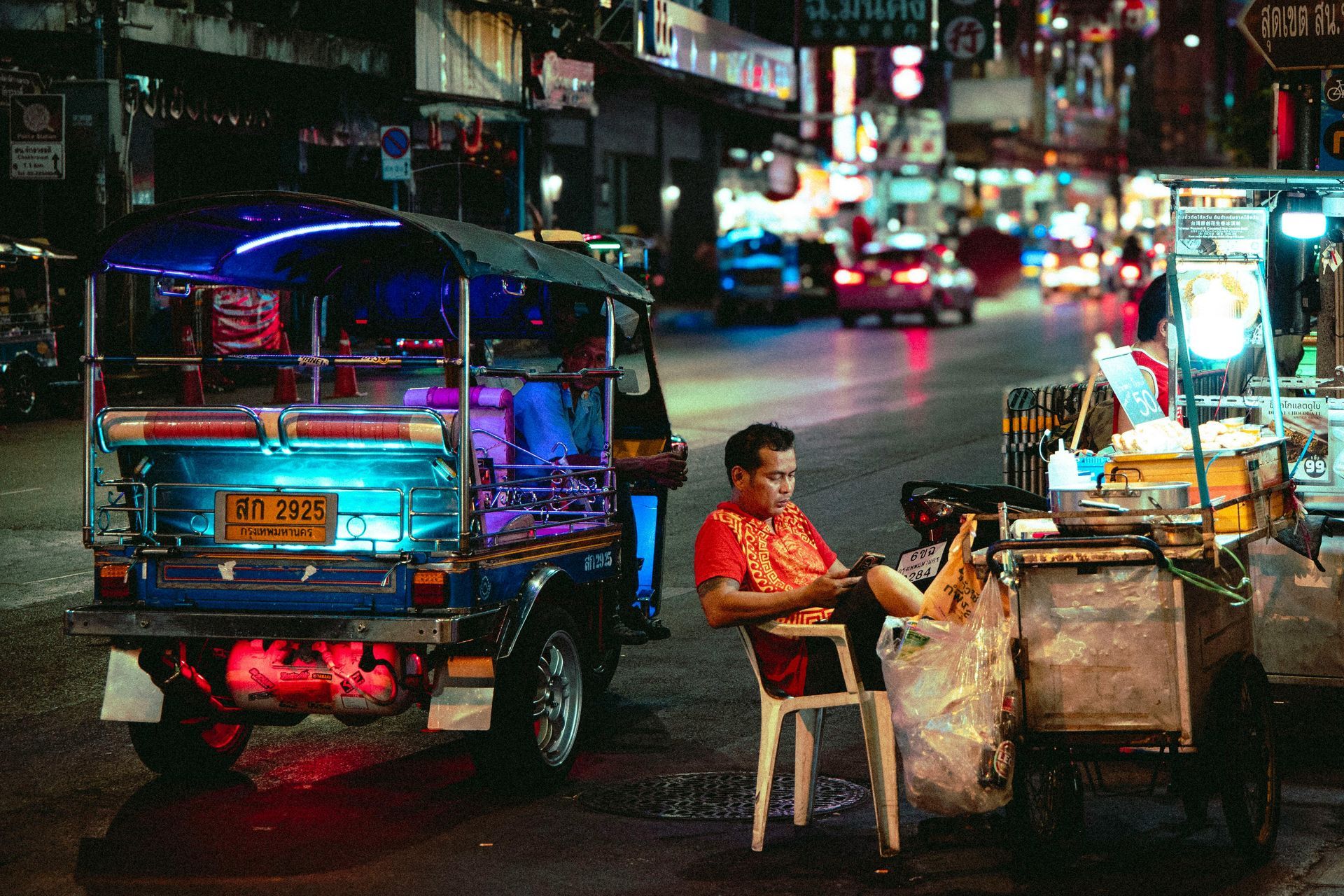 Night street scene with a brightly lit tuk-tuk and a vendor sitting beside a food cart.