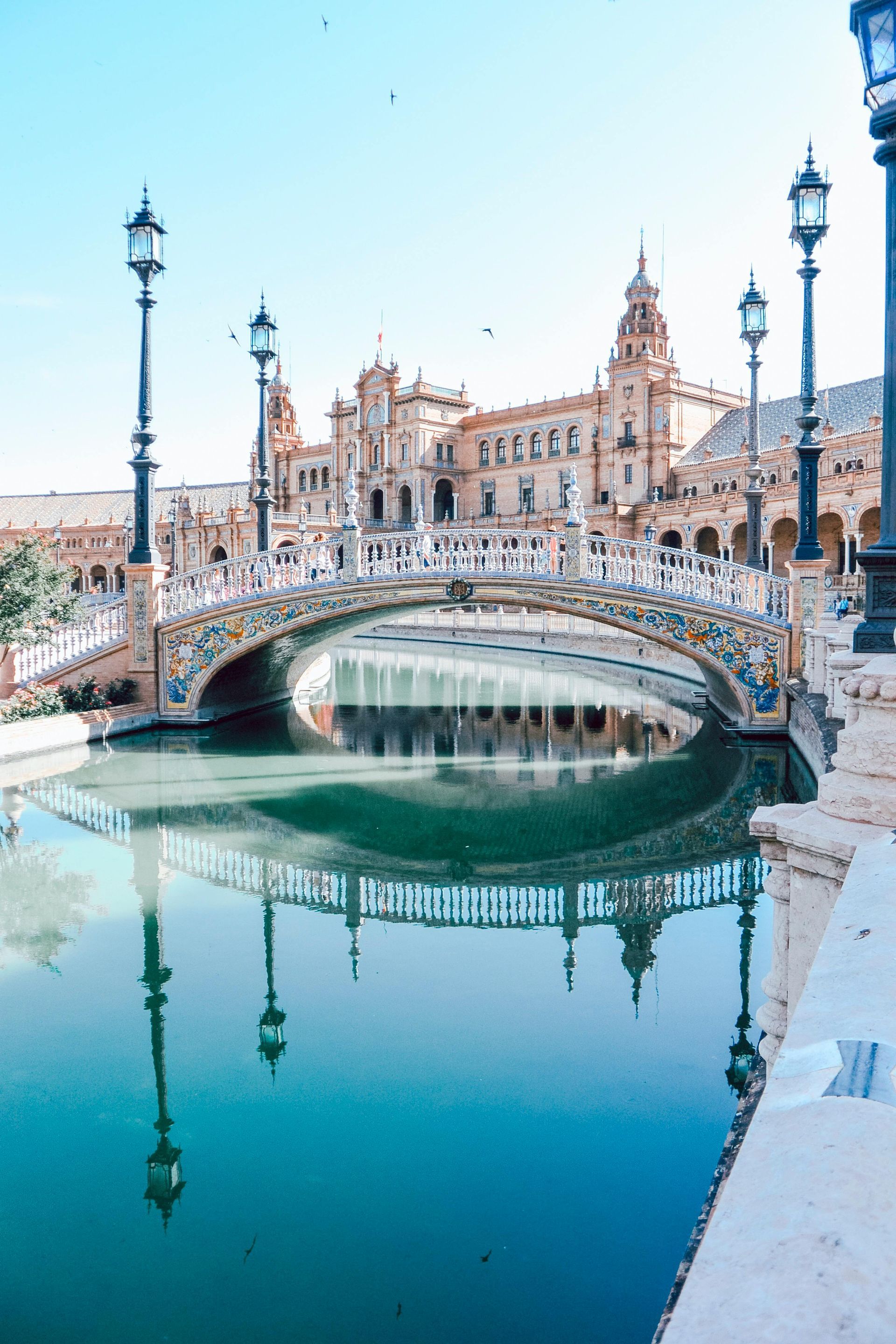 Canal with arched bridge and ornate buildings reflected in calm turquoise water, framed by street lamps