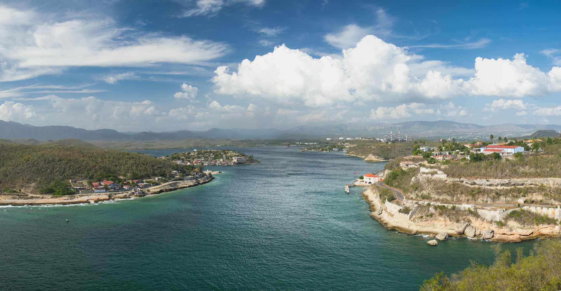 Wide turquoise bay between rocky cliffs under a bright, cloudy sky, with a distant shoreline