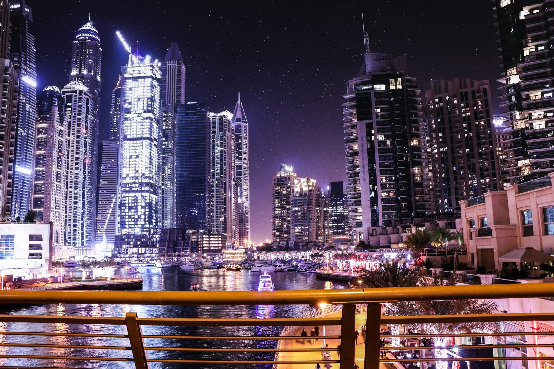 Nighttime city skyline with illuminated skyscrapers and waterfront reflections, seen through a railing