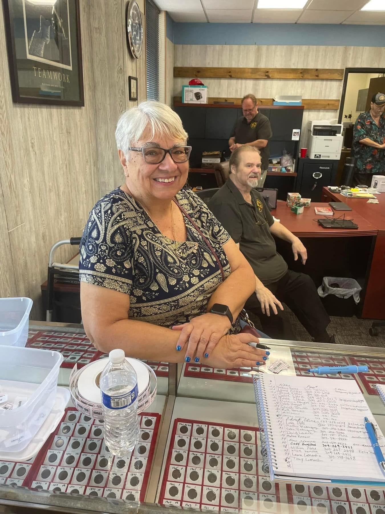 Smiling woman at a desk with displayed coin and stamp collections in a shop office