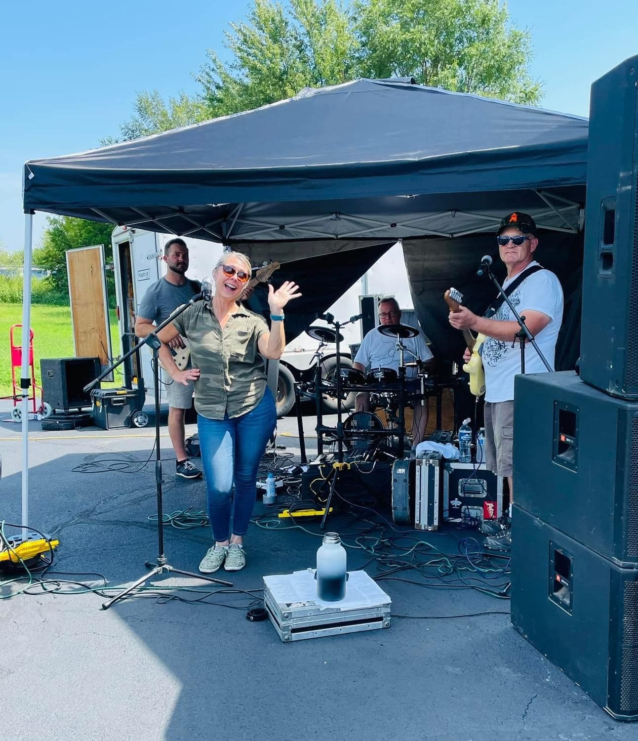 Outdoor band performing under a black canopy, with singer at front and drummer and guitarist behind.