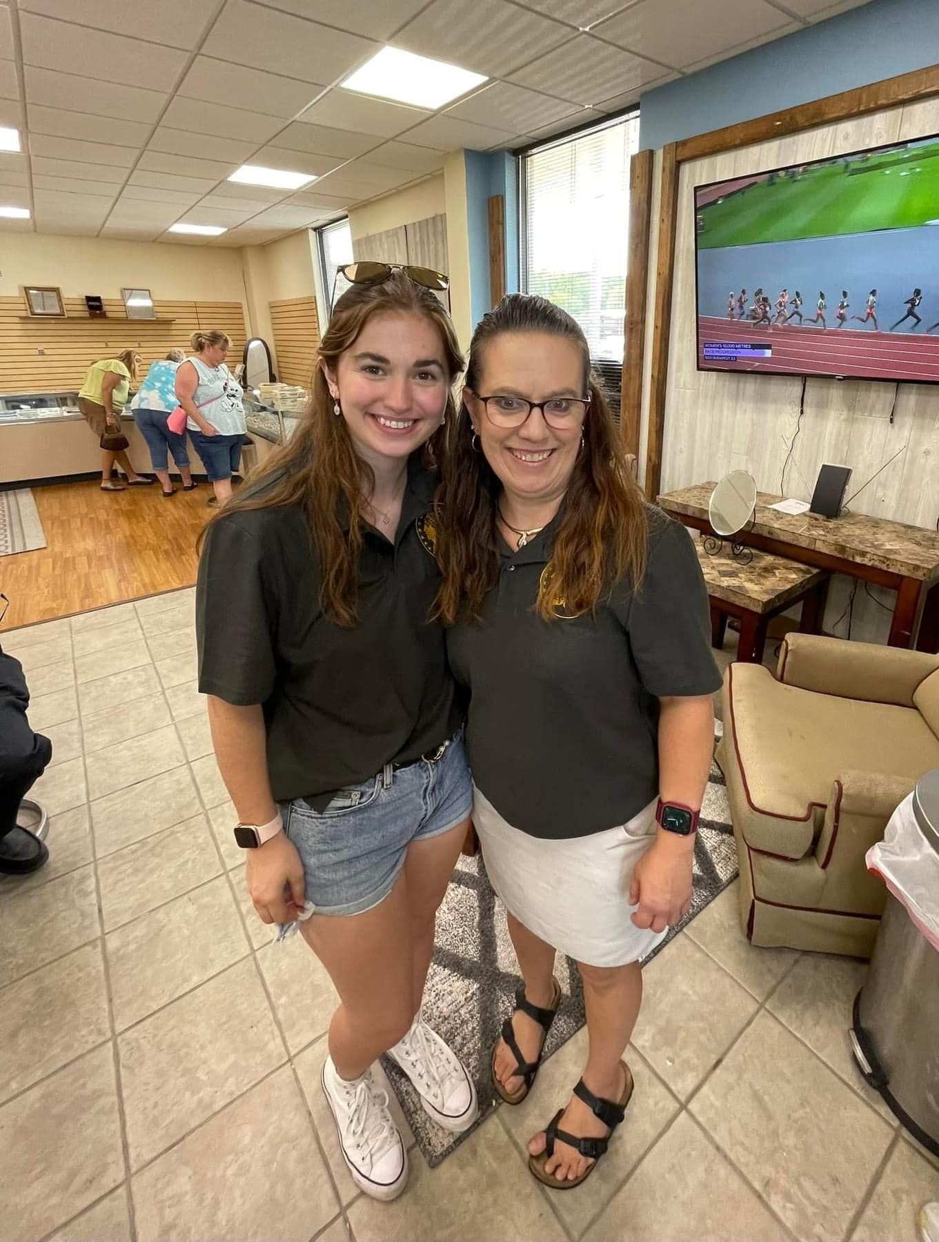 Two smiling women pose in a lounge, with chairs and a wall-mounted TV in the background.