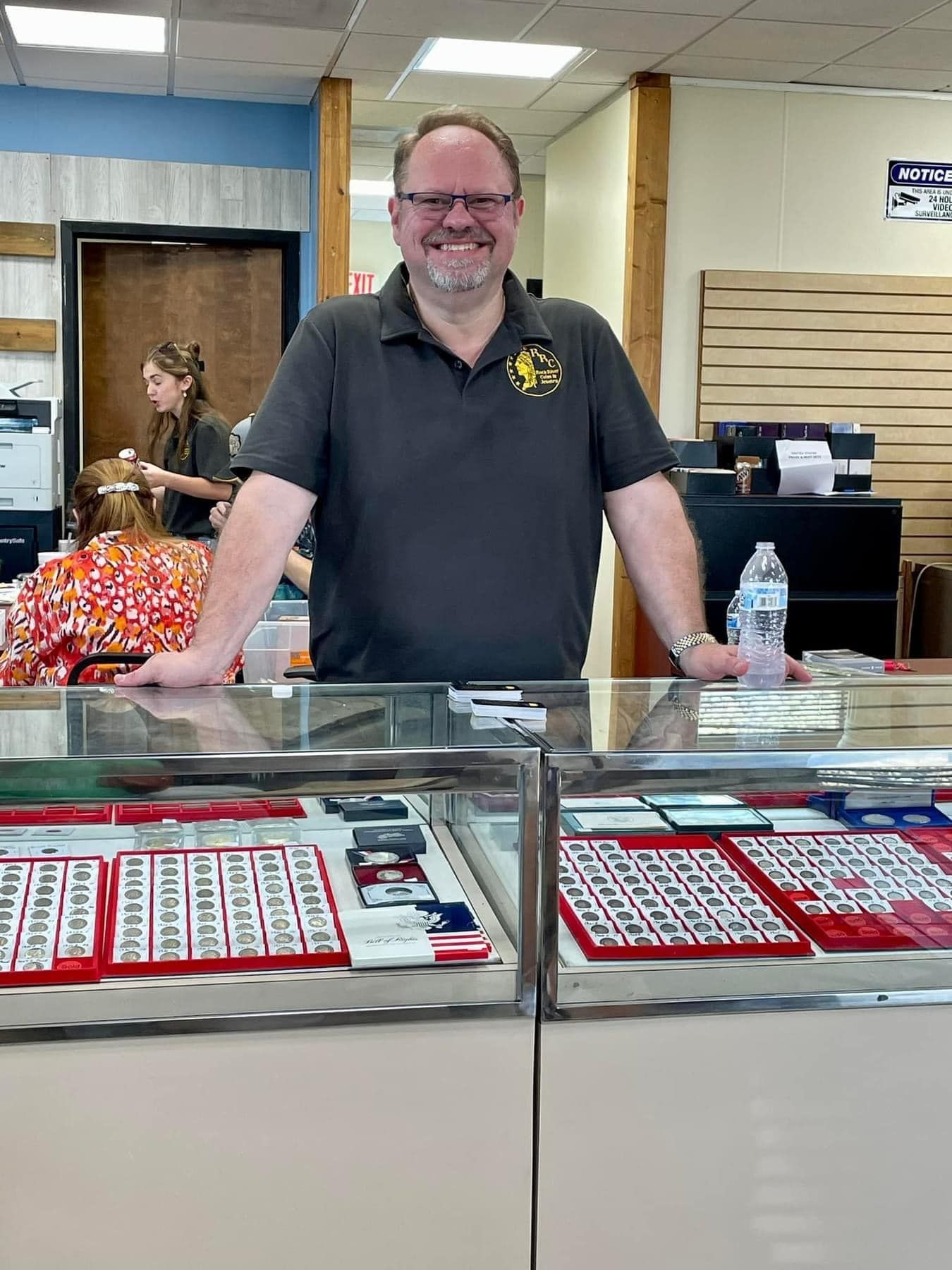 Smiling man behind a jewelry display counter in a store, with red trays and a water bottle.