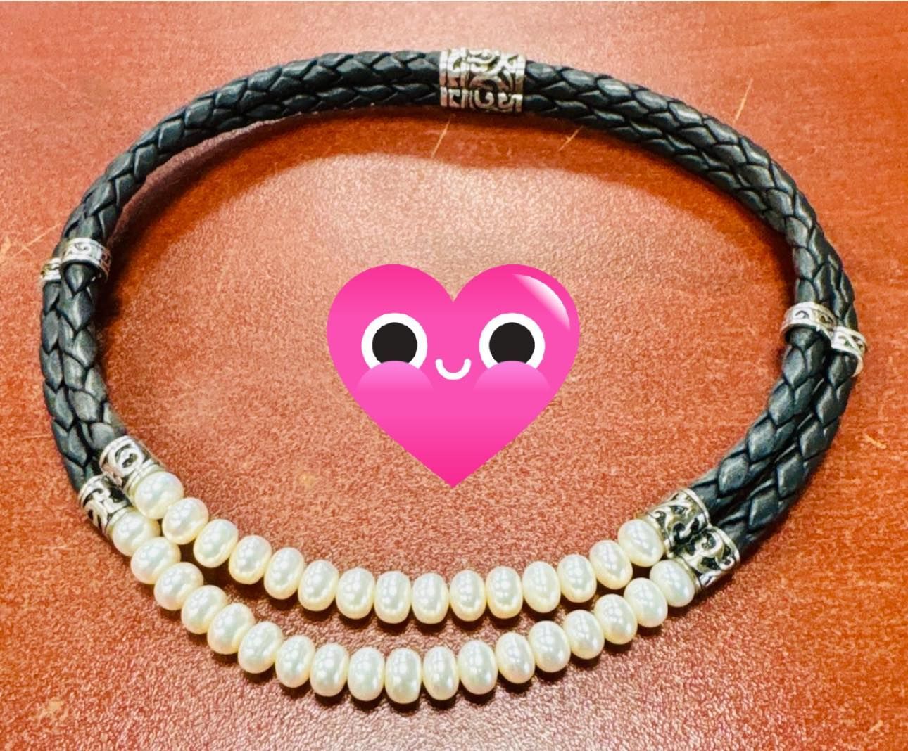 Black braided necklace with two rows of pearls on a brown background, centered with a pink heart emoji