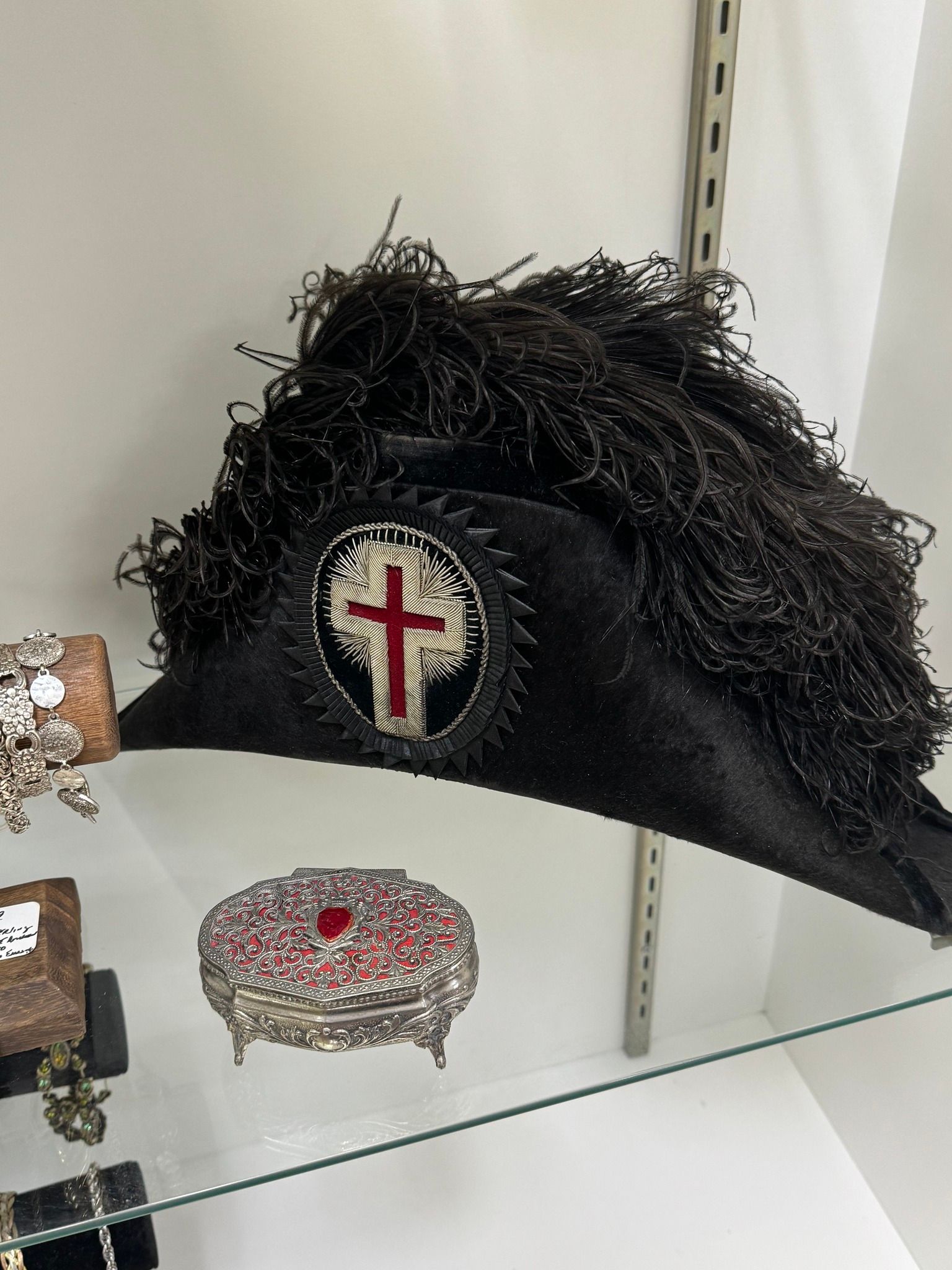 Black military-style hat with a furry plume and cross emblem on a glass shelf beside a jeweled brooch.