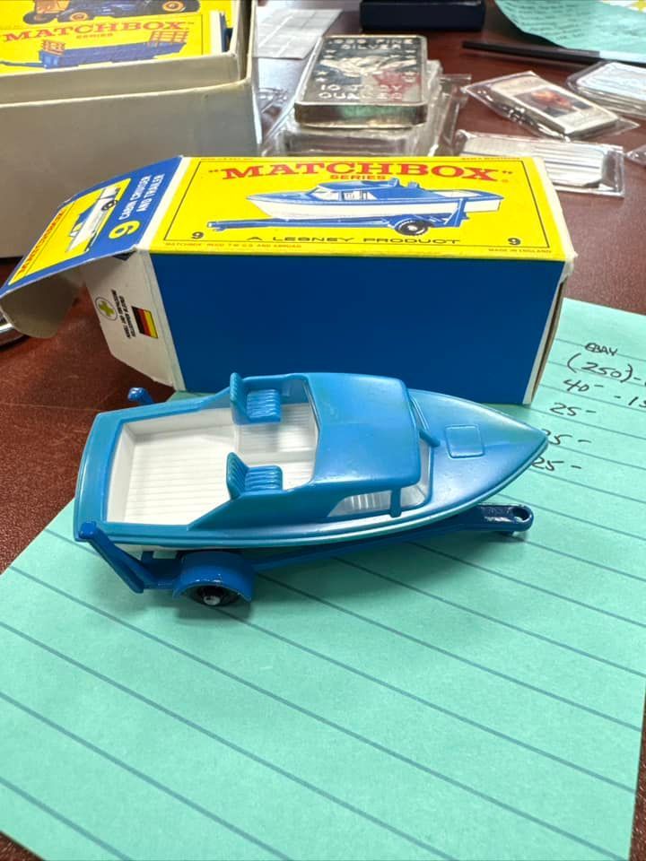 Blue toy speedboat with white seats on a desk, beside a yellow Matchbox box and papers.