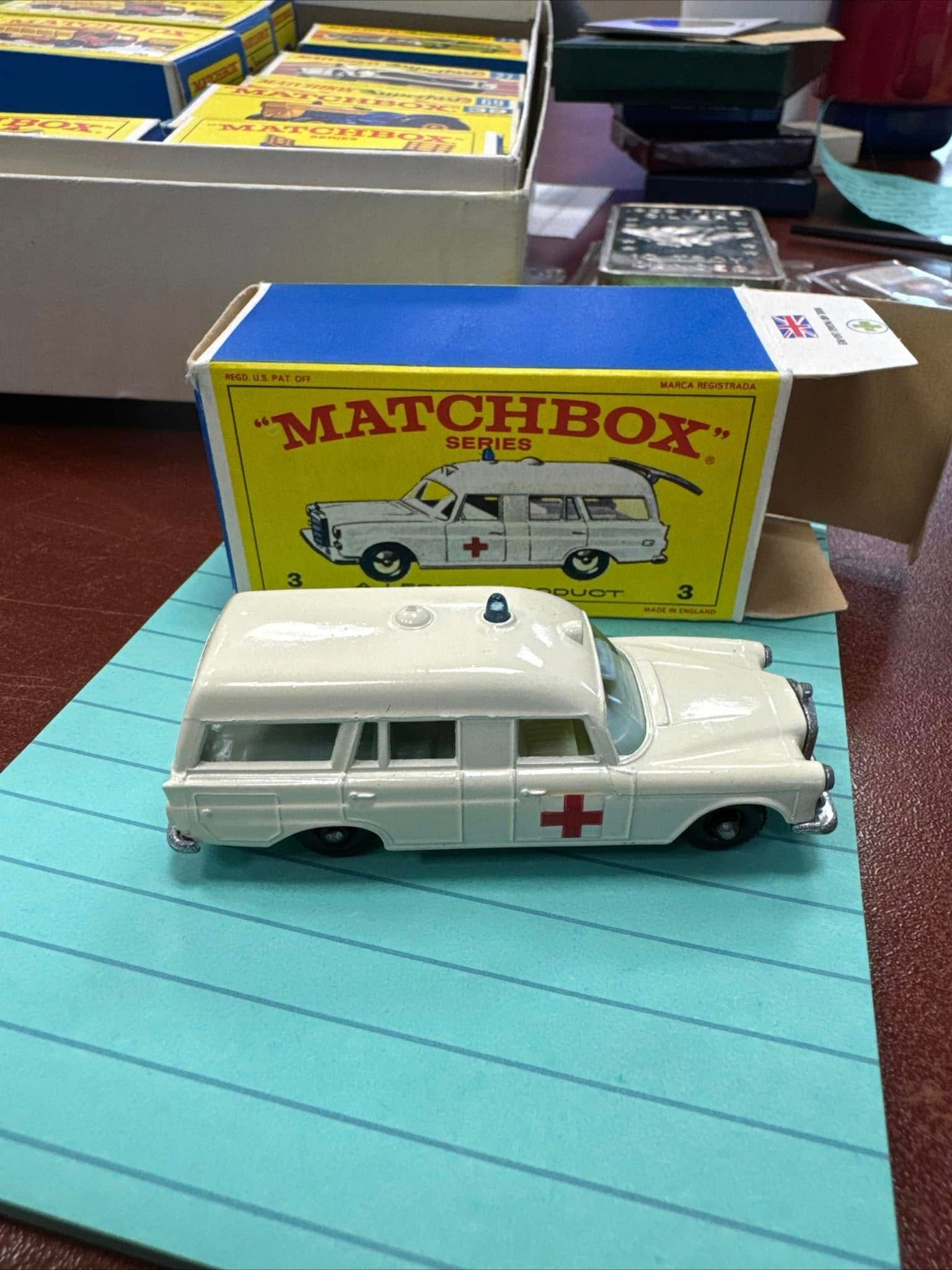 White Matchbox toy ambulance with red crosses on a blue table beside its box.