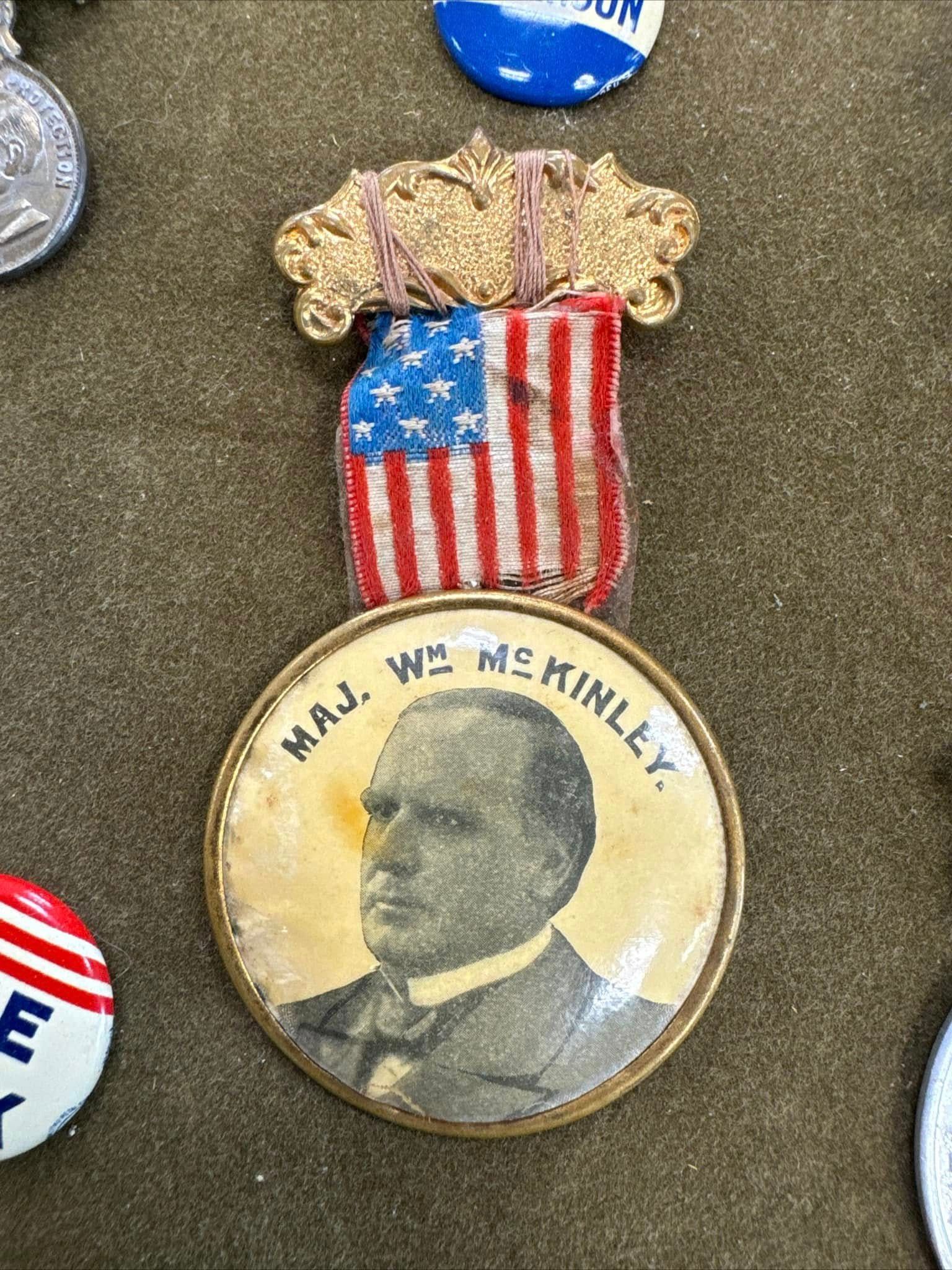 Round pin with a portrait and text, topped by a small American flag emblem.