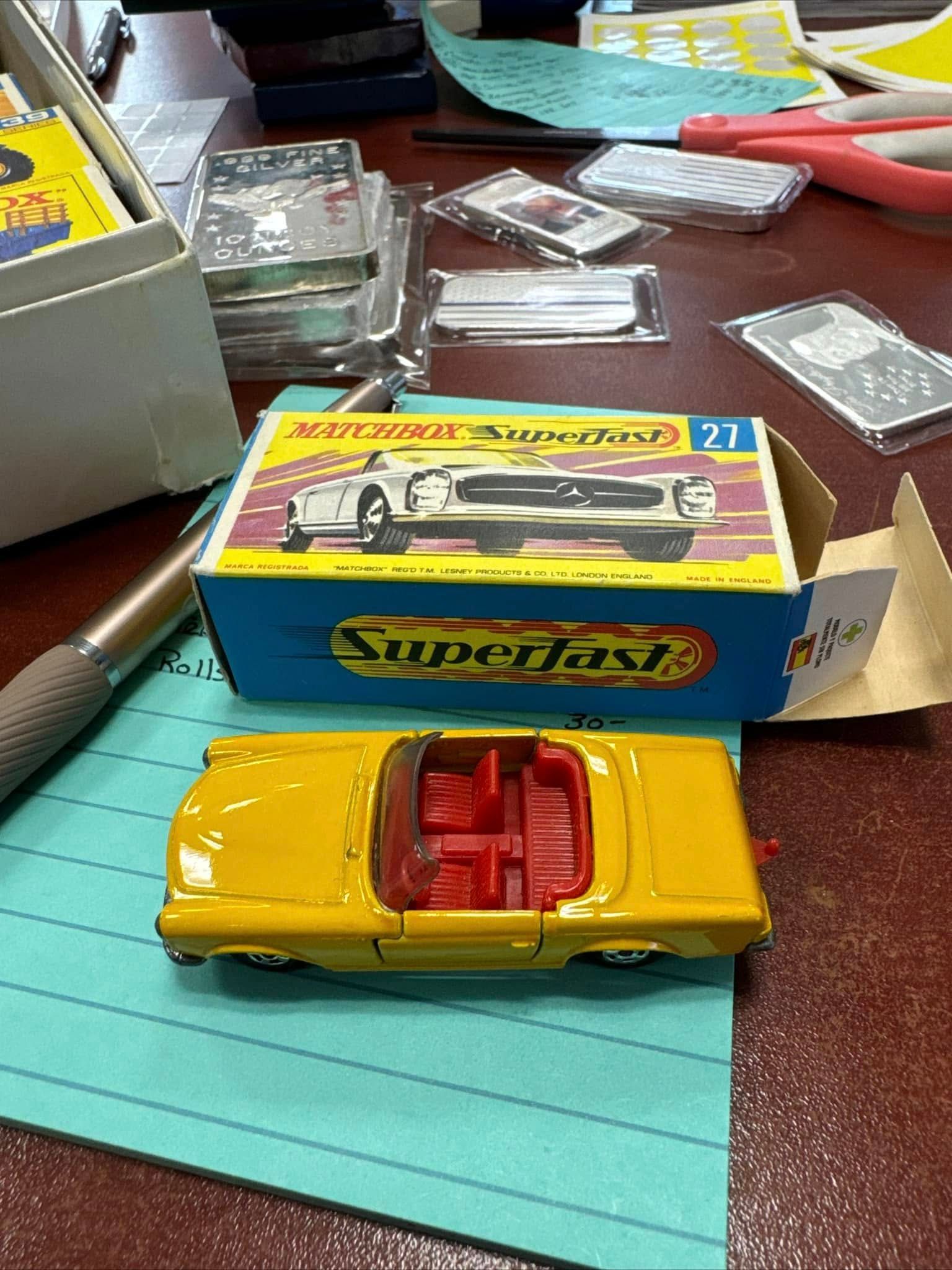 Yellow toy car on a desk in front of a Matchbox box, with small clear plastic cases nearby.