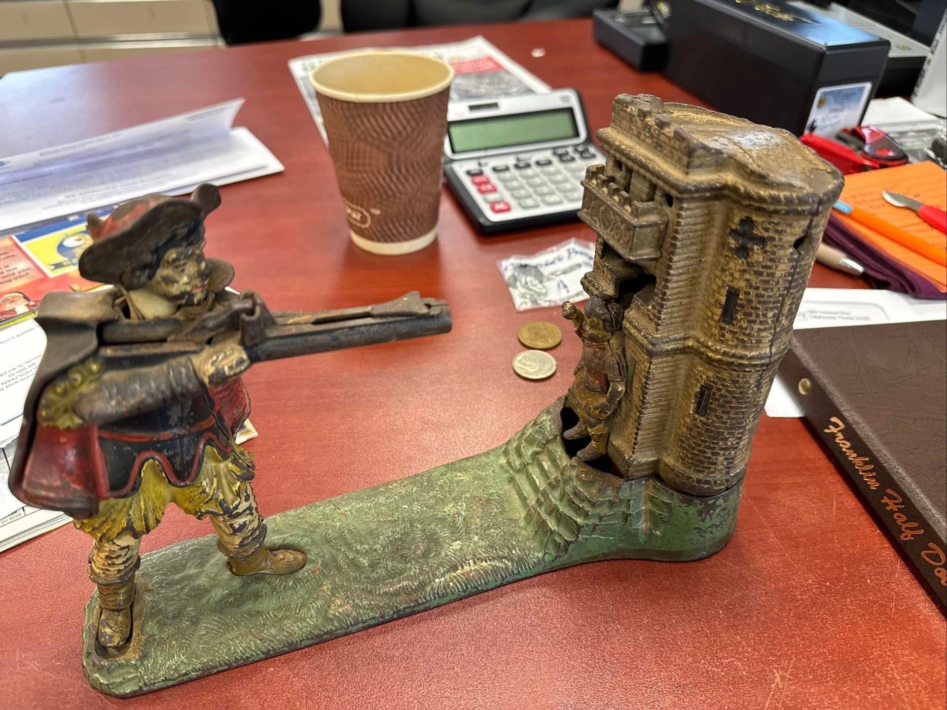 Small figurine beside a crumbling tower model on a desk with coins, calculator, and a paper cup.