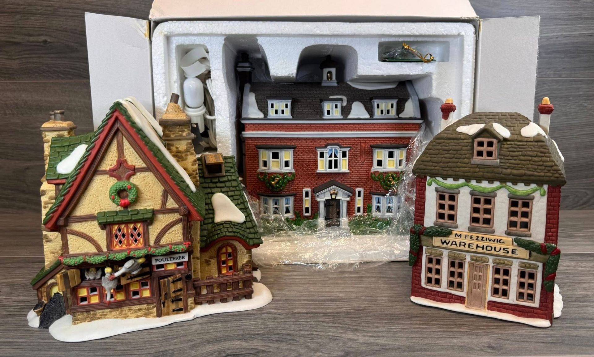 Miniature Christmas village houses with lit storefronts and snow on the rooftops, displayed in a box.