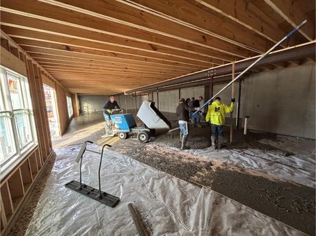Construction workers pouring concrete inside a building with wooden beams, using a dumper and leveling tools.