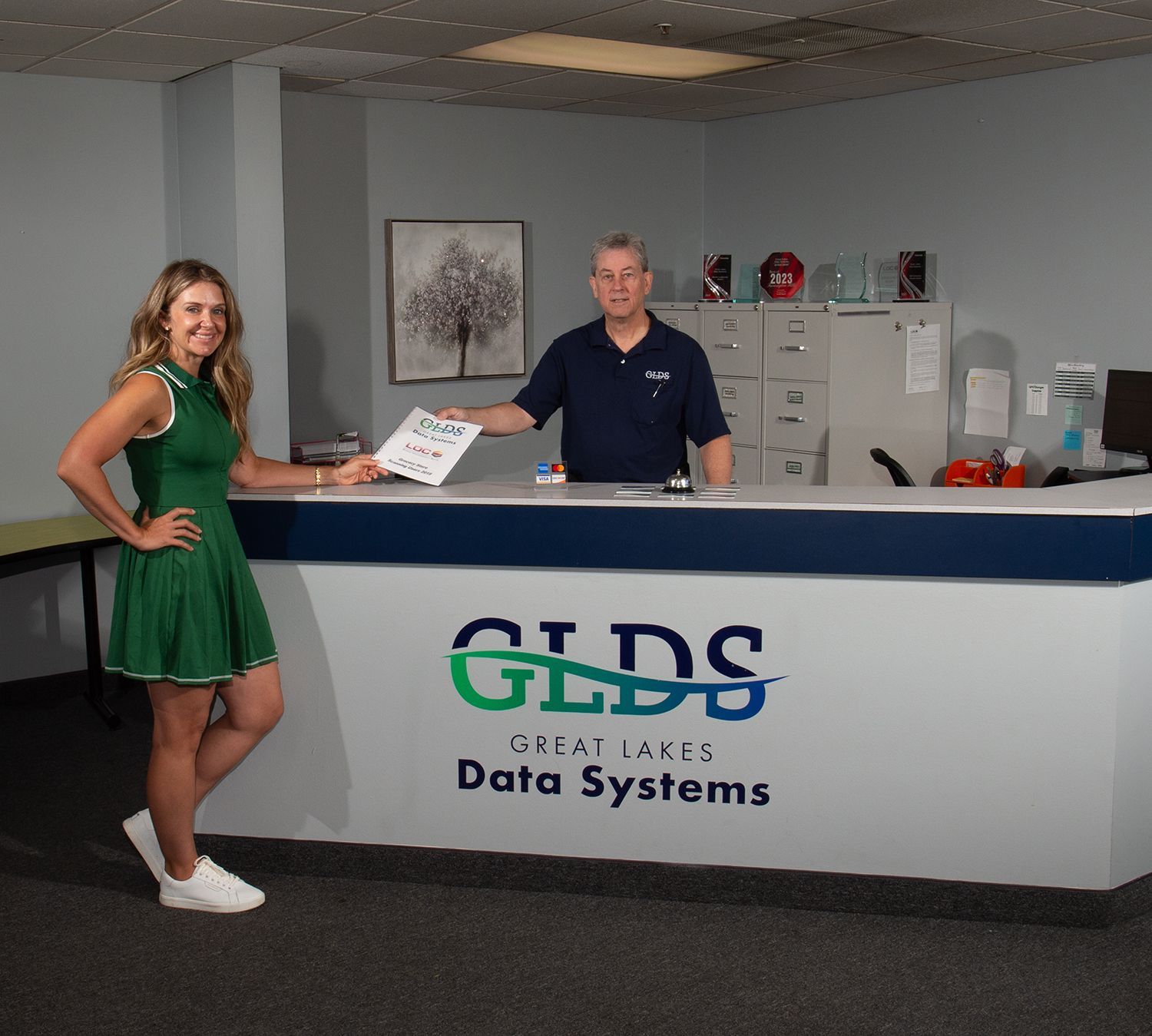 Two people stand behind a reception desk with GLDS Great Lakes Data Systems logo in an office lobby.