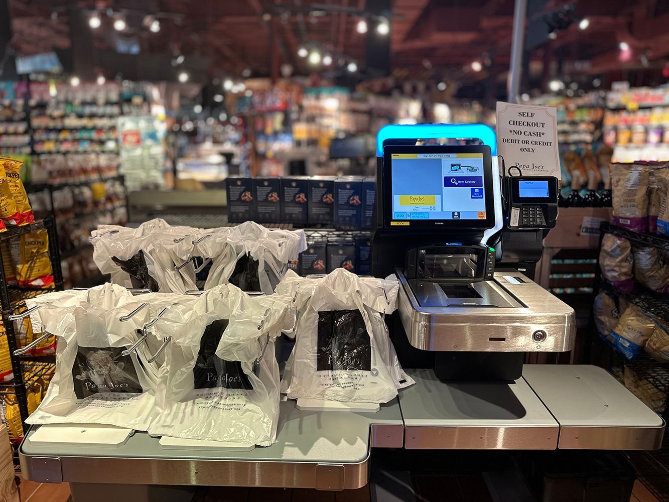 Grocery checkout counter with bagged items and a glowing self-checkout screen in a store.